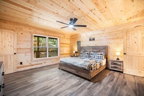 Une chambre dans une cabane en bois, avec un lit, un ventilateur de plafond, une fenêtre et une table de chevet.