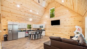 Une pièce spacieuse avec un plafond en bois, un coin repas comprenant une table et des chaises, un canapé et une cuisine dotée de meubles blancs et d’un réfrigérateur.