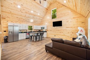 Ein geräumiger Raum mit Holzbalkendecke, einem Essbereich mit Tisch und Stühlen, einer Couch und einer Küche mit weißen Schränken und einem Kühlschrank.