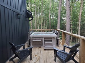 A hot tub on a wooden deck with two chairs, surrounded by trees.