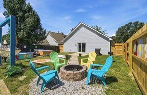 Ein Garten mit Feuerstelle, bunten Adirondack-Stühlen und einem Holzzaun.