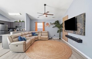 A modern living room with a sectional sofa, a flat-screen TV mounted on the wall, a ceiling fan, and a kitchen in the background. - Cowboy pool Hot tub Boho Charlotte (Charlotte)