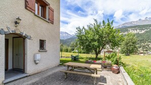 A building with a stone facade, a wooden door, and a window with shutters. There is a wooden bench and a table outside, with a view of a tree and mountains in the background. - Androsace - VAL CENIS (VAL CENIS)