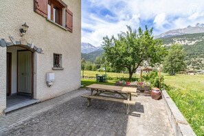 Ein Gebäude mit einer steinernen Fassade, einer Holztür und einem Fenster mit Fensterläden. Vor der Tür stehen eine hölzerne Bank und ein Tisch, mit Blick auf einen Baum und Berge im Hintergrund.