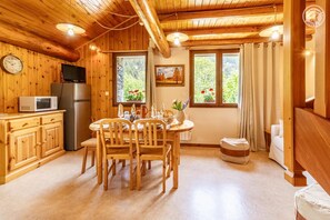 Ein Holzhütten-Interieur mit Esstisch, Stühlen, einer Küchenzeile mit Geräten und einem großen Fenster mit Vorhängen.