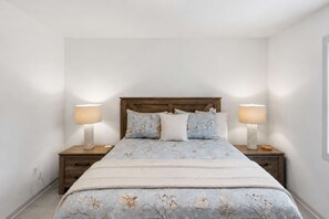 A bedroom with a wooden headboard, floral bedding, two bedside lamps, and a nightstand with a candle. - New- Eastpoint- Next to RV park (Eastpoint)