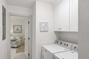 A laundry room with a washer and dryer, white cabinets, and a door leading to a living area with a couch and a rug. - New- Eastpoint- Next to RV park (Eastpoint)