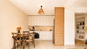 Una cocina compacta con armarios blancos, una mesa de comedor con sillas, una lámpara colgante y un separador tejido.