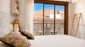 Un dormitorio con cama, mesita de noche, lámpara, ventana con vista a un edificio y balcón con sillas y una mesa.