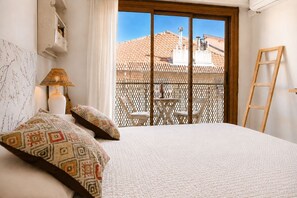 Ein Schlafzimmer mit einem Bett, einem Nachttisch, einer Lampe, einem Fenster mit Blick auf ein Gebäude und einem Balkon mit Stühlen und einem Tisch.
