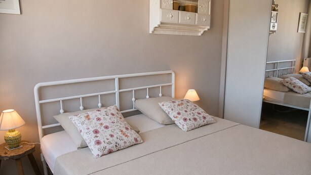 Una cama con estructura metálica blanca, dos almohadas estampadas con motivos florales, un edredón color beige y una lámpara de noche sobre una mesita de madera.