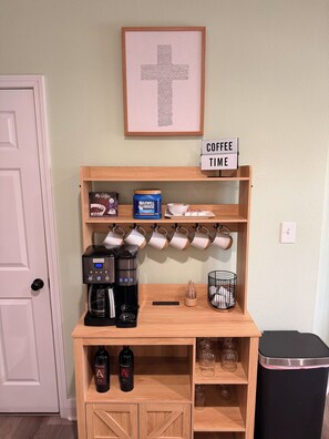 Un coin café avec un panneau indiquant l’heure du café, une machine à café, des tasses, un récipient à café et des bouteilles de vin.