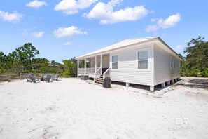 Ein eingeschossiges Haus mit Veranda, gelegen an einem sandigen Strand, mit Bäumen im Hintergrund.