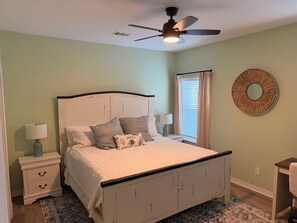 A bedroom with a bed, nightstands, a ceiling fan, a window with curtains, and a wall-mounted circular mirror. - The Lou | Walk 2 Beach, Golf Cart & Beach Toys! (Gulf Shores)