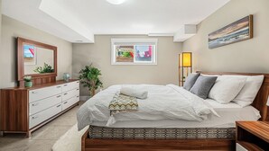 A bedroom with a bed, dresser, bedside table, lamp, and a window with a view. - Family Basement Retreat Near Rockies (Okotoks)