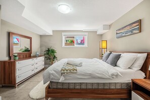 Ein Schlafzimmer mit Bett, Kommode, Nachttisch, Lampe und einem Fenster mit Blick.