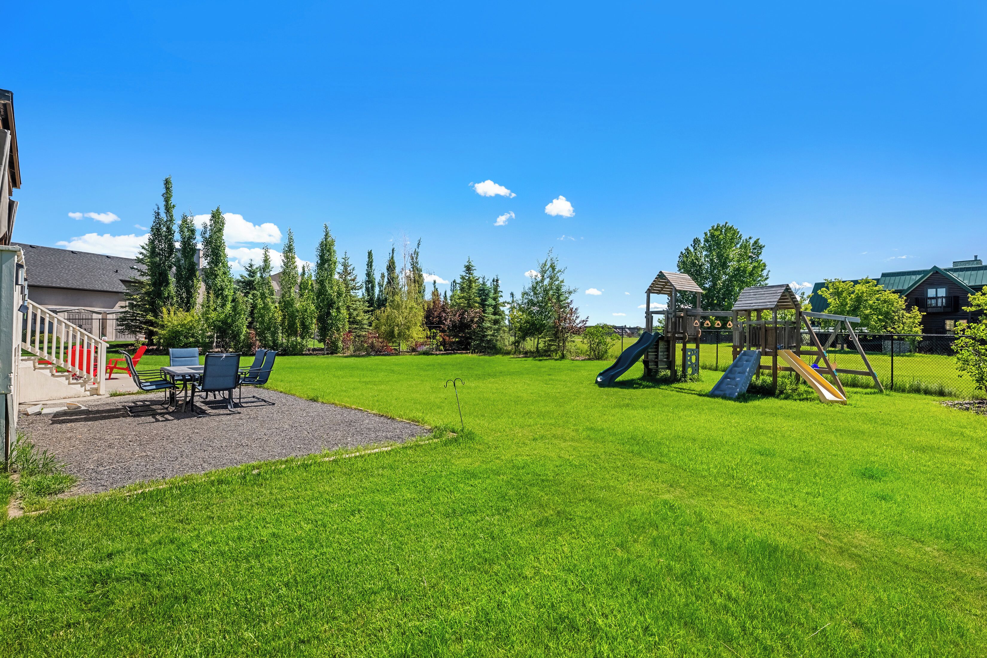 A grassy backyard with a playground, a patio with chairs, and a building with a staircase.