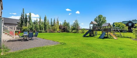 A grassy backyard with a playground, a patio with chairs, and a building with a staircase.