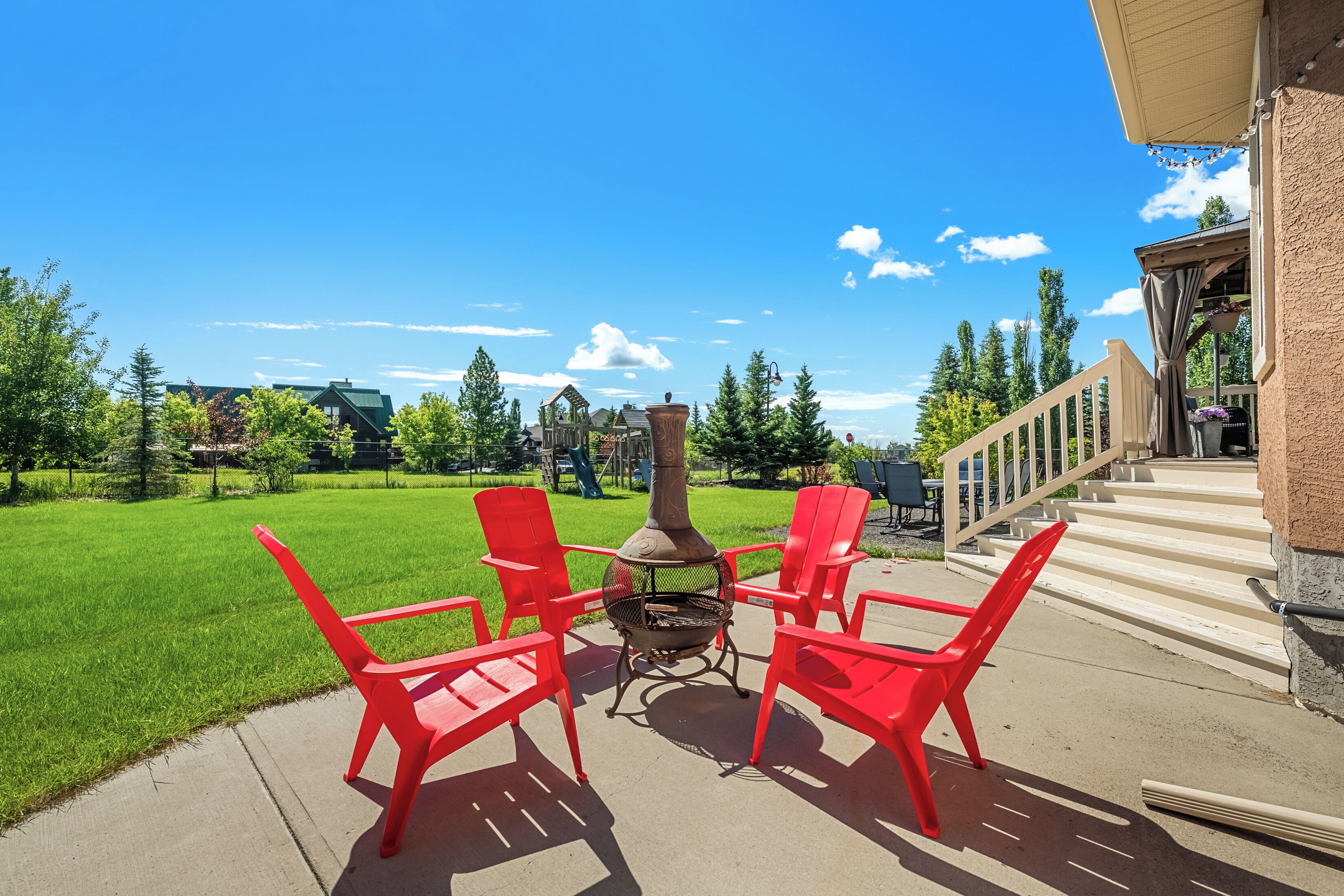 Une terrasse avec des chaises rouges et un foyer, donnant sur une pelouse verdoyante et une aire de jeux.