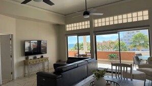 A spacious living area with a flat-screen TV, a sectional sofa, a dining table with a potted plant, and large windows offering a view of the ocean and buildings. - A302 Casa Blanca Golf Villas 3-bedroom condo with AC in Puerto Peñasco (Puerto Peñasco)