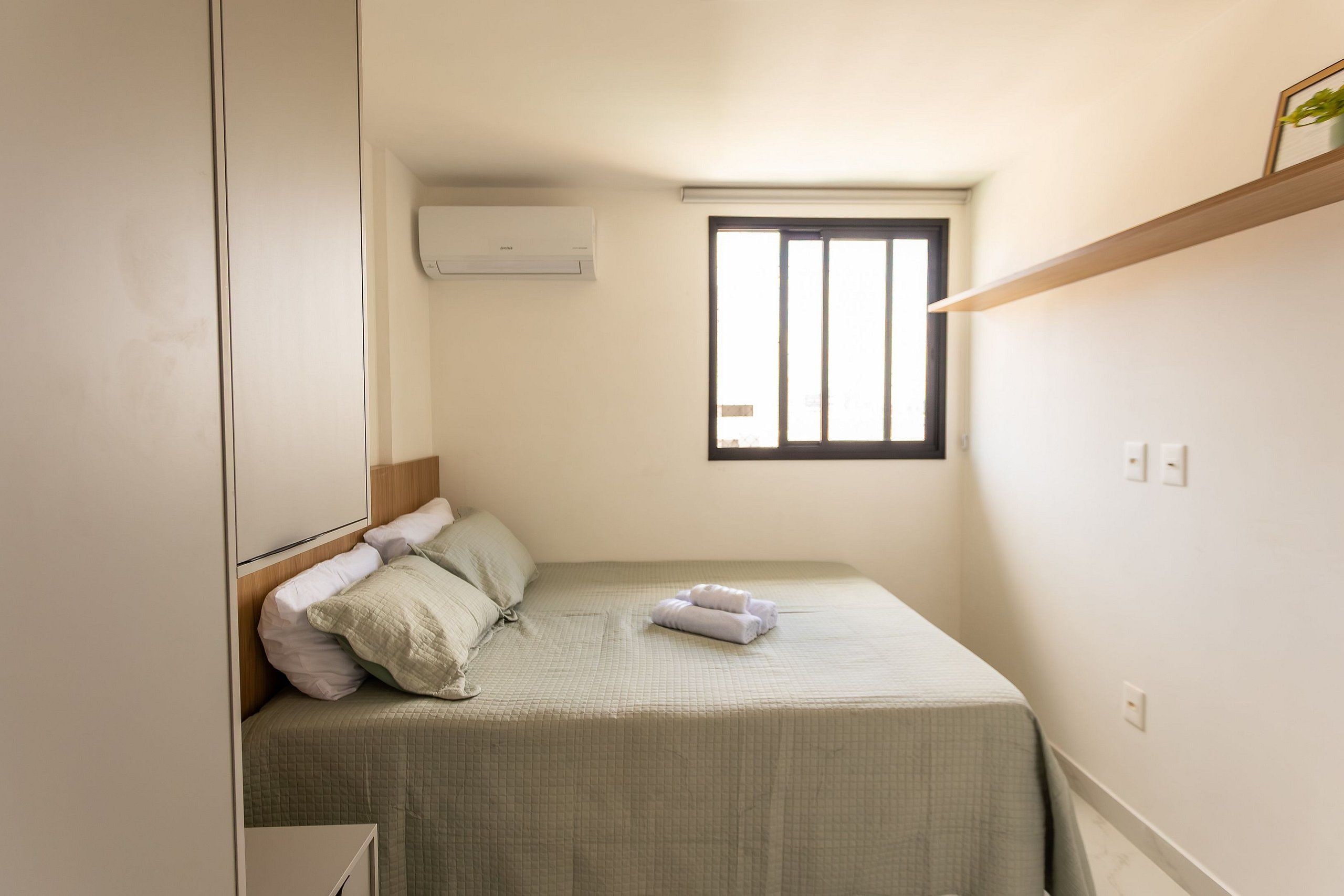 Un dormitorio pequeño y ordenado con una cama individual, una ventana con rejas, un aire acondicionado de pared y un estante sobre la cama.