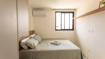 Un dormitorio pequeño y ordenado con una cama individual, una ventana con rejas, un aire acondicionado de pared y un estante sobre la cama.