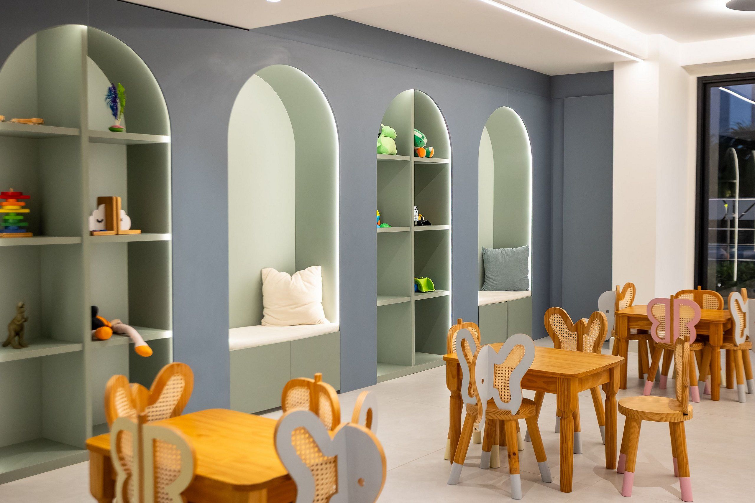A children's playroom with arched shelving, wooden tables, and chairs.