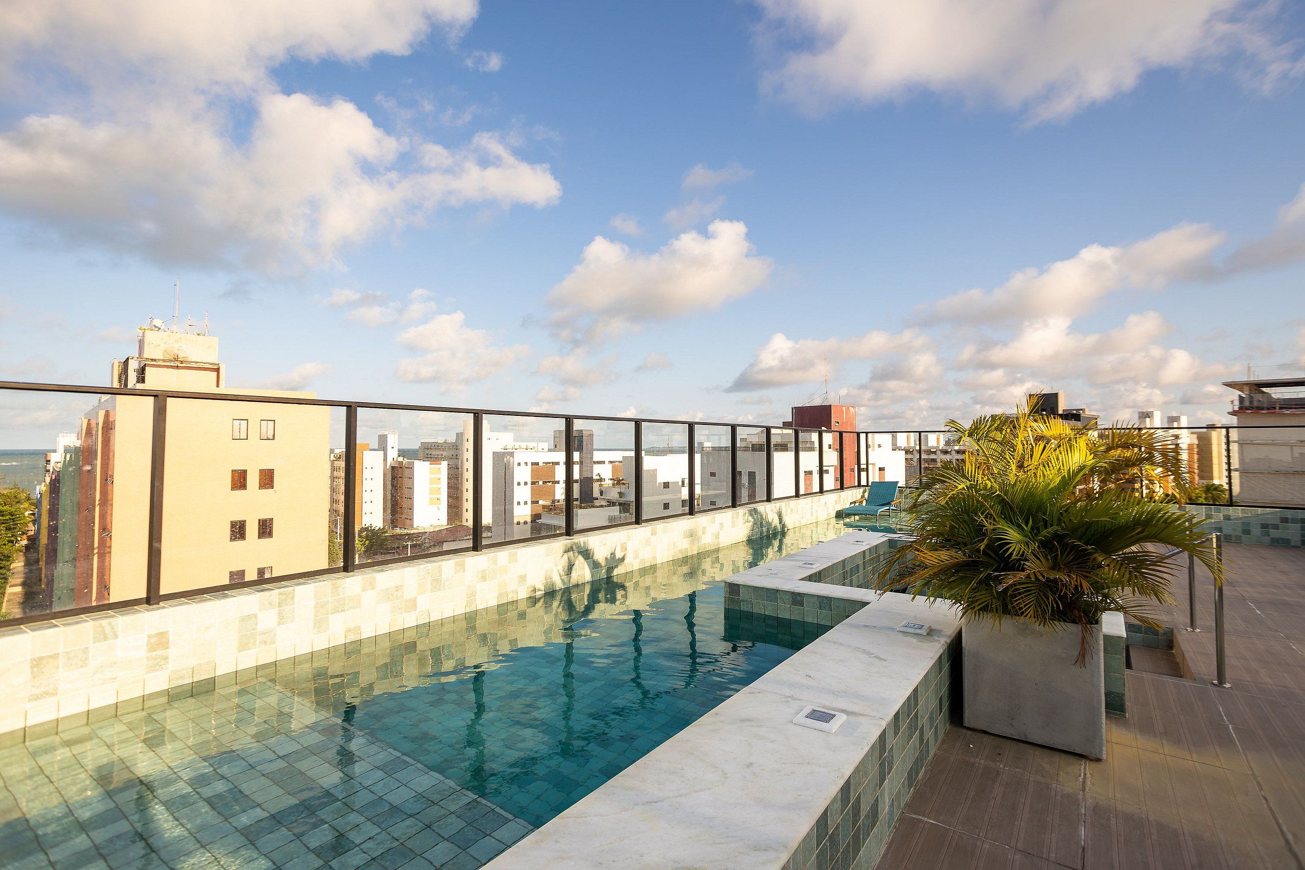 Piscina no terraço com água azul cristalina, cercada por guarda-corpo de vidro, com vista para um horizonte urbano com diversos edifícios.