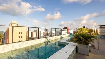 Piscina no terraço com água azul cristalina, cercada por guarda-corpo de vidro, com vista para um horizonte urbano com diversos edifícios.