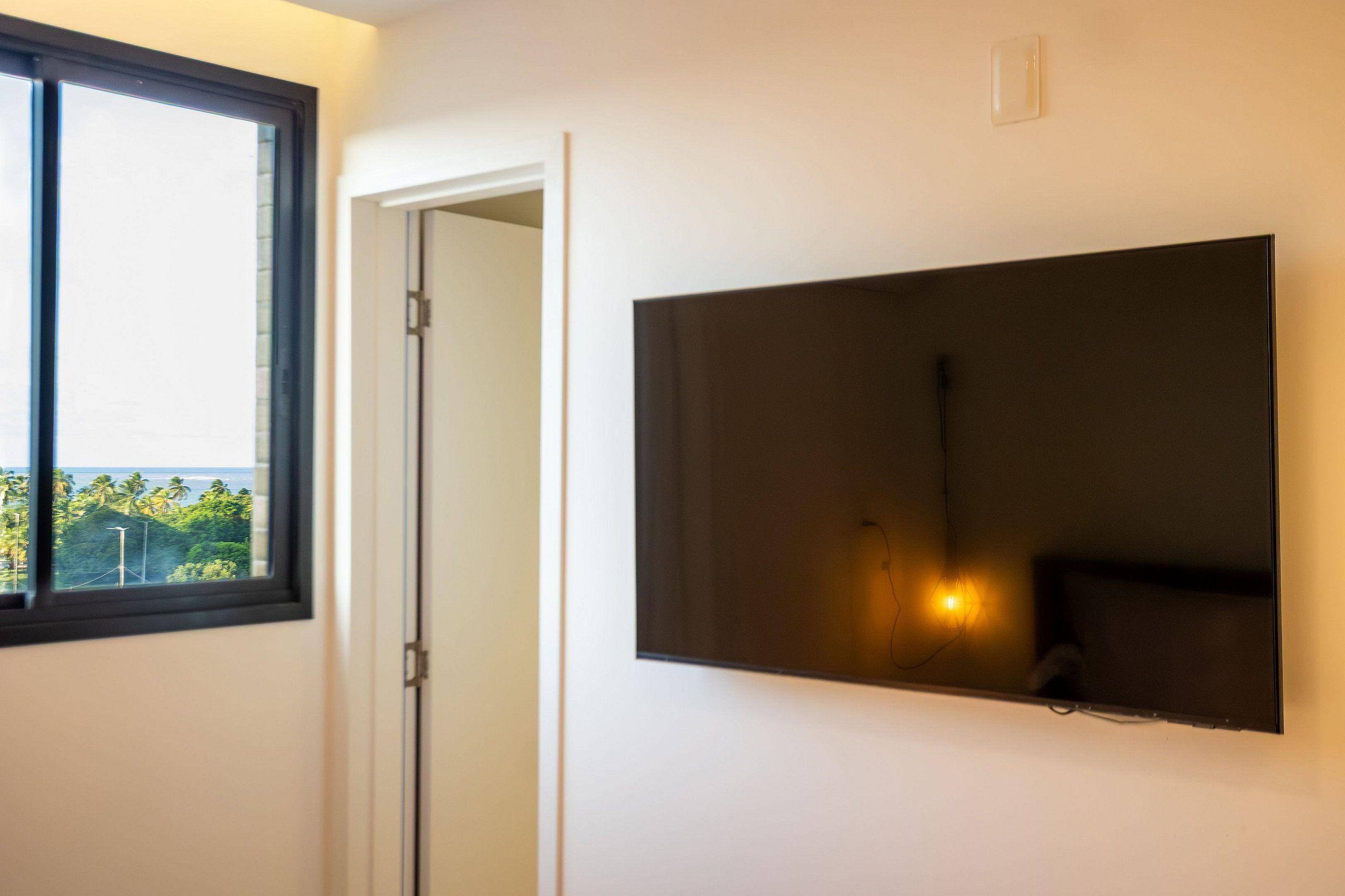 Una habitación con un televisor de pantalla plana grande montado en la pared, una ventana con vistas a zonas verdes, y una puerta que da a otra habitación.