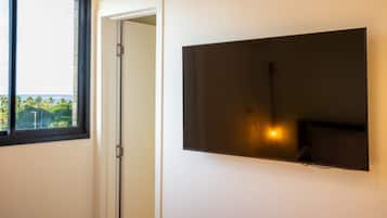 Una habitación con un televisor de pantalla plana grande montado en la pared, una ventana con vistas a zonas verdes, y una puerta que da a otra habitación.