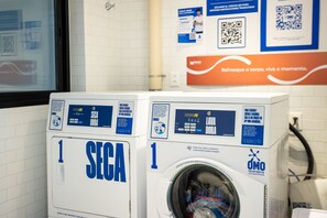 Two front-loading washing machines, one labeled 'SEGA' and the other 'OMO,' in a laundry room with tiled walls and a poster above them. - NOA Residence Club in Ponta de Campina With Rooftop and Full Leisure by Carpediem (Cabedelo)