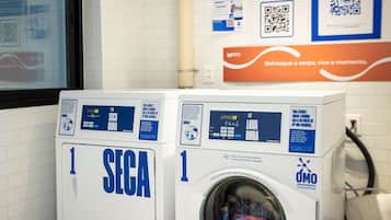 Two front-loading washing machines, one labeled 'SEGA' and the other 'OMO,' in a laundry room with tiled walls and a poster above them.