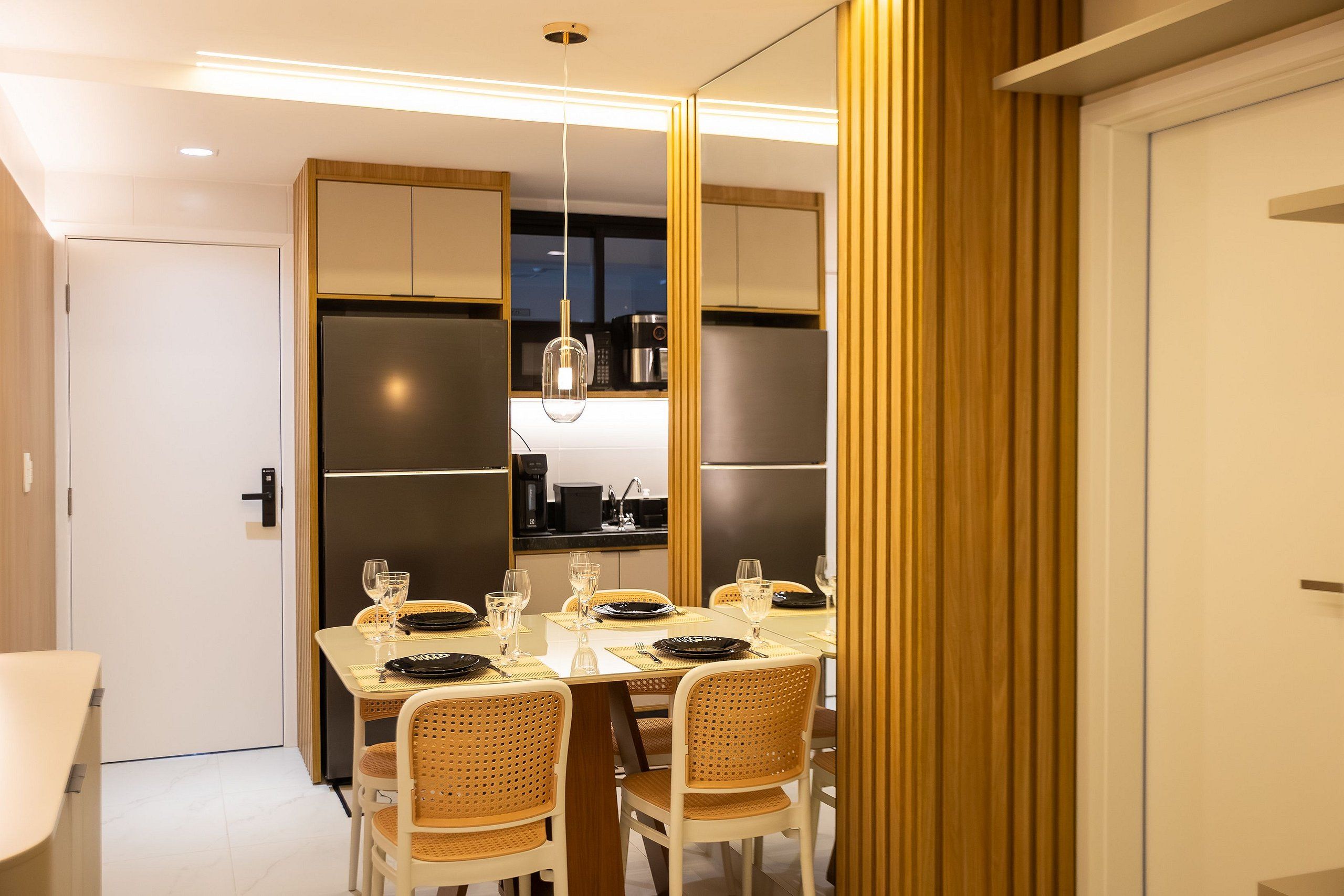 Uma área de refeições moderna com uma mesa posta para quatro pessoas, rodeada por cadeiras de vime, e uma cozinha visível através de uma divisória.
