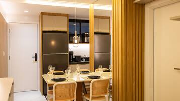 Uma área de refeições moderna com uma mesa posta para quatro pessoas, rodeada por cadeiras de vime, e uma cozinha visível através de uma divisória.