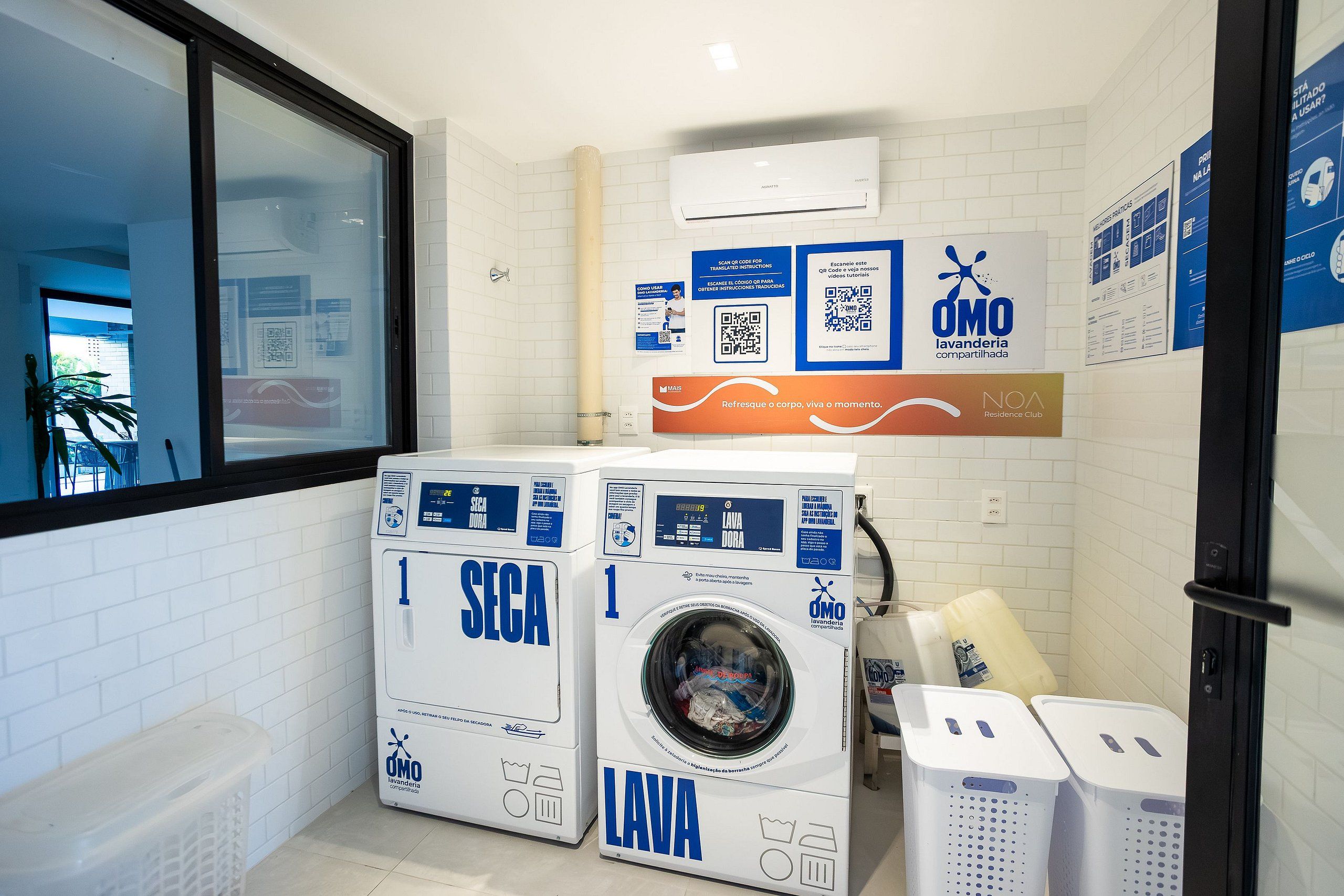 Un área de lavandería con dos lavadoras, un aire acondicionado de pared y carteles informativos.
