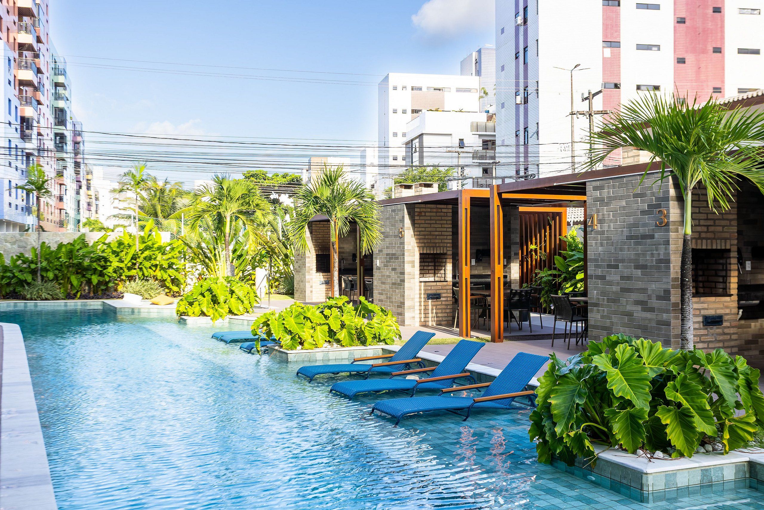 A rooftop pool with blue lounge chairs, surrounded by greenery and modern buildings.