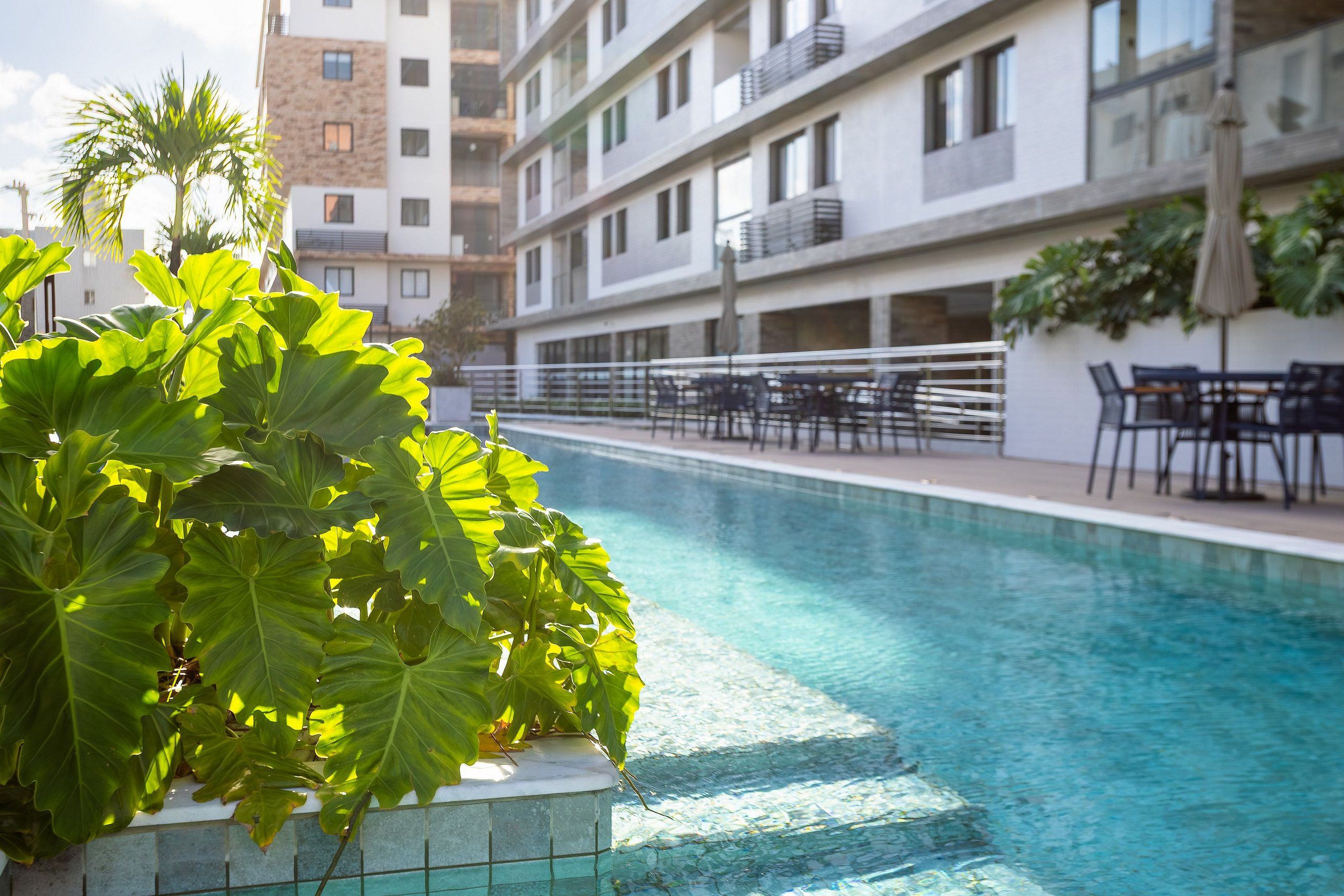 A swimming pool with a tiled edge, surrounded by outdoor seating and a building with multiple balconies in the background.