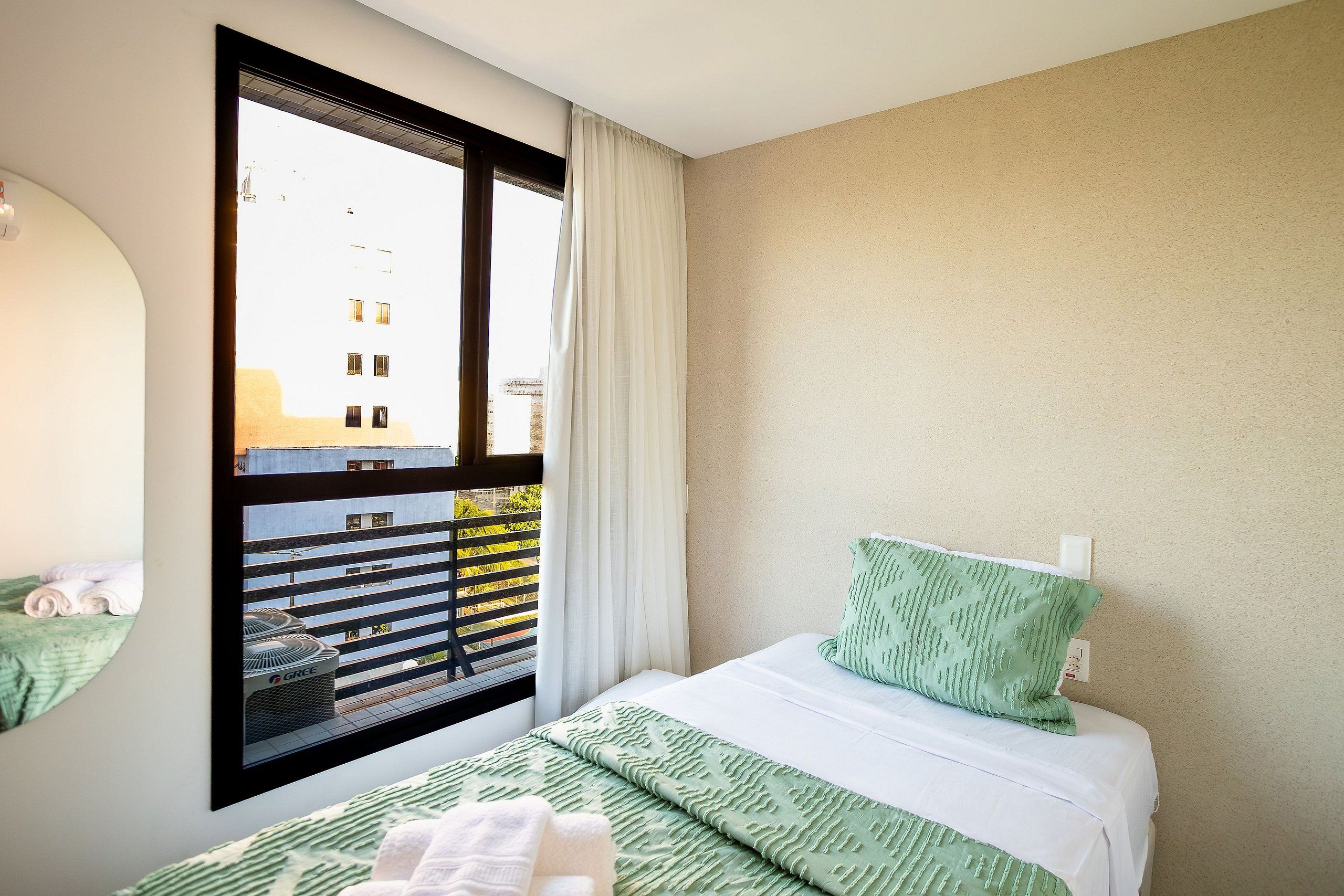 Habitación de hotel con un ventanal grande, una cama con ropa de cama verde, almohada blanca y vistas a edificios.