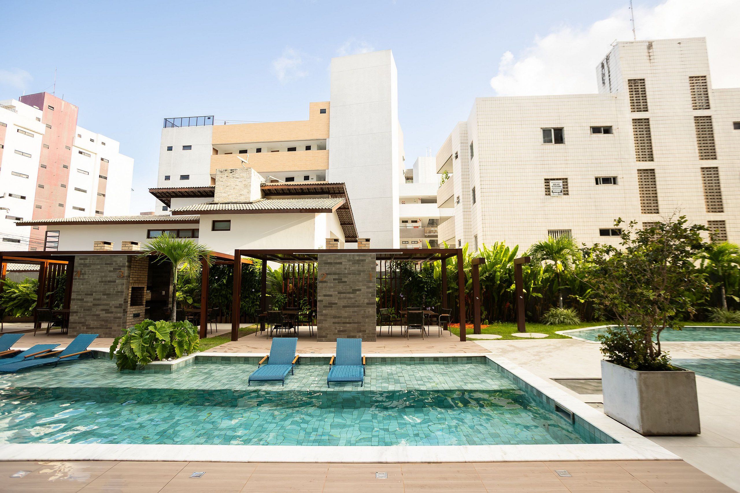 Una piscina en la azotea con sillones de descanso y una pérgola, rodeada de vegetación y edificios modernos.