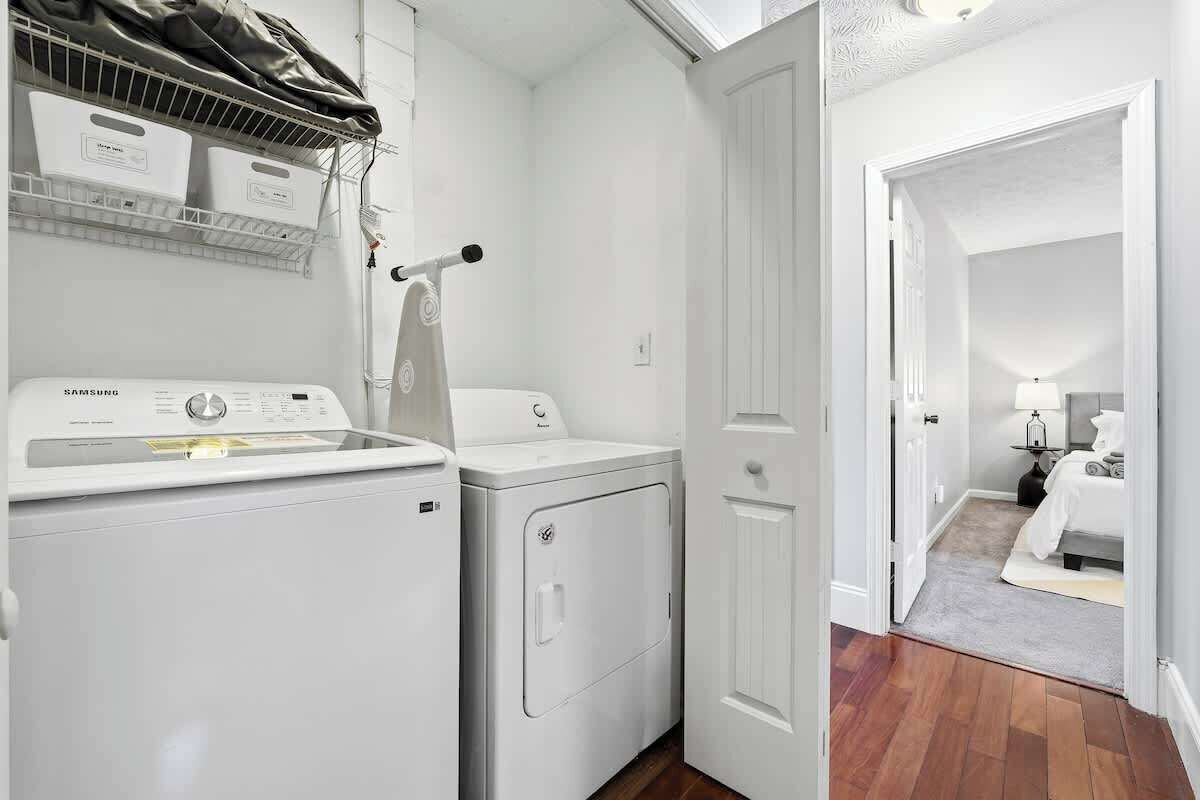 A laundry area with a Samsung washer and dryer, a drying rack, and a door leading to a bedroom with a bed and nightstand.