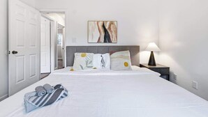 Hotellihuone, jossa on sänky, tyynyjä, yöpöytä, lamppu ja taulu seinällä.