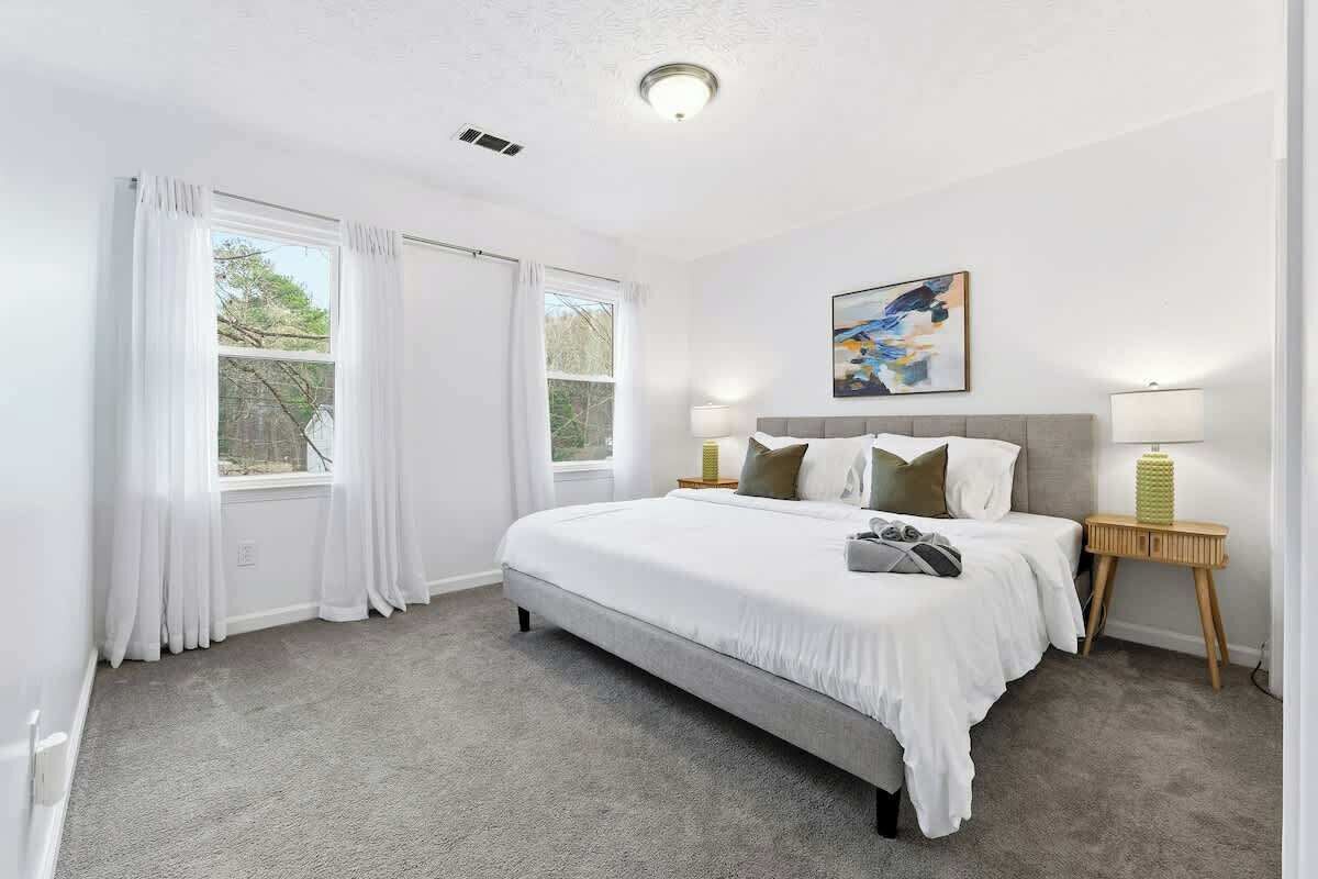 A bedroom with a bed, two bedside tables, a window with curtains, a painting on the wall, and a ceiling light.