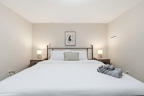 A hotel room with a large bed, two bedside lamps, a framed artwork on the wall, and a neatly made bed with towels. - Windsor House | All-King| Sun-Filled Game Room (Lawrenceville)