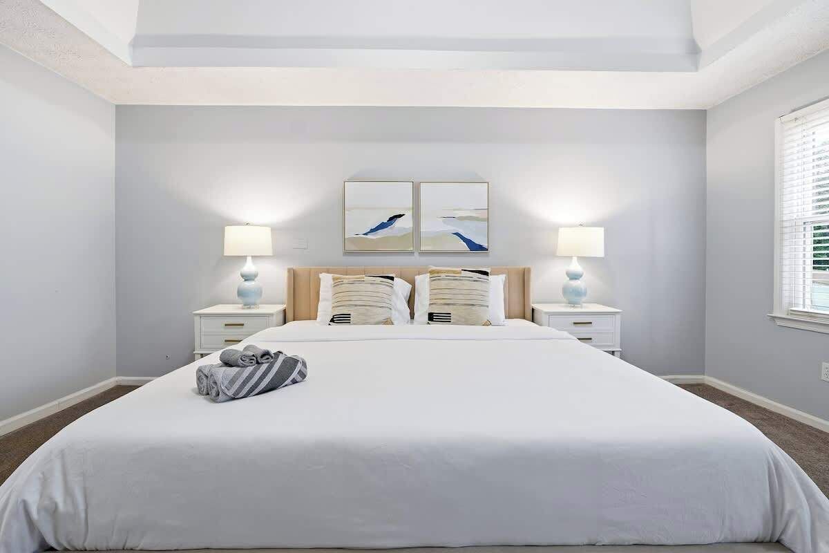 A bedroom with a large bed, two bedside tables with lamps, a headboard with artwork, and a window with blinds.