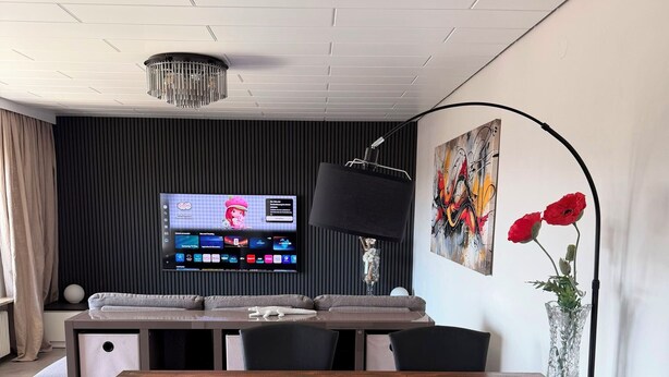 Un comedor moderno con una mesa de madera, sillas negras, un televisor de pantalla plana, una lámpara de pie negra y un jarrón con flores rojas.