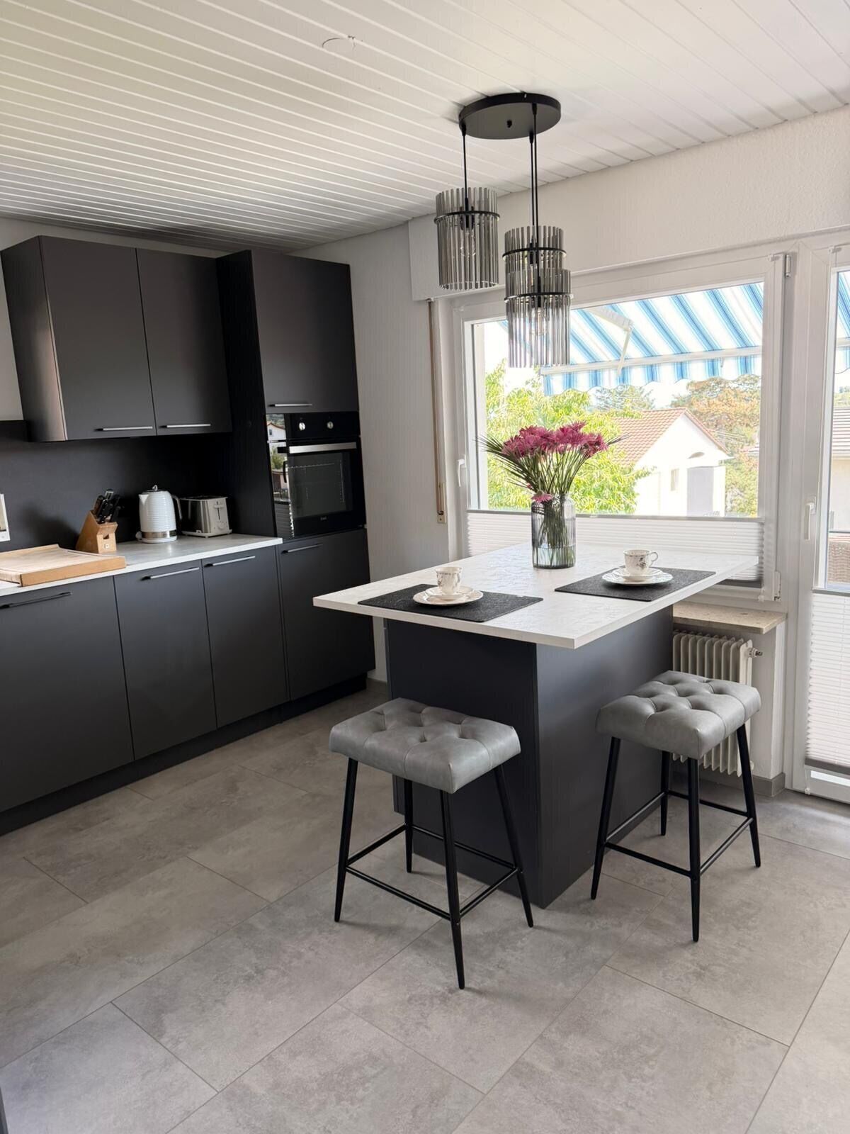 A modern kitchen with dark cabinets, a central island, bar stools, and a window with a view.