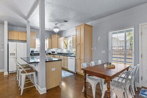A modern kitchen with wooden cabinets, a central island, and a dining area with white chairs. - 3BR Coastal Retreat | 10 Min to Beaches (Pensacola)