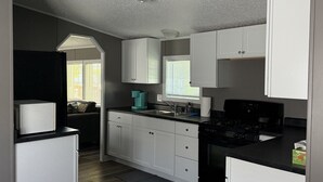 A compact kitchen with white cabinets, a black stove, and a window with blinds. - Peaceful 3-Bedroom home on Lake Oahe (Pierre)