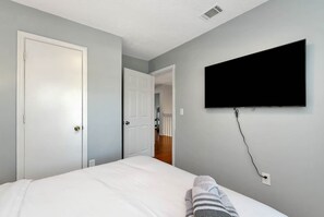 A bedroom with a bed, a TV mounted on the wall, and a door leading to another room. - Daylilly House/ Family Friendly with Gameroom (Lawrenceville)